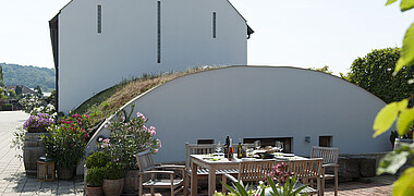 Weißes Haus mit Giebeldach und halbrunden Weinkeller-Gewölbe davor in einem grünen Garten.