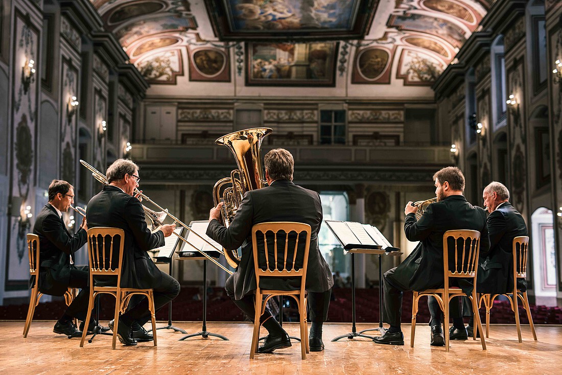 Fünf Männer mit Blechblasinstrumenten auf einer Bühne in einem großen barocken Konzertsaal.
