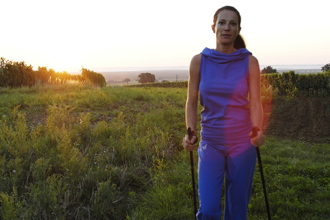 Frau in lila Trainingsbekleidung geht mit Nordic Walking Stöcken durch Weingärten