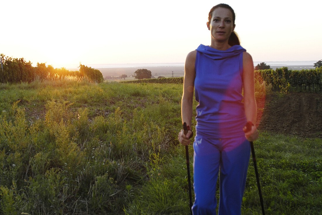 Frau in lila Trainingsbekleidung geht mit Nordic Walking Stöcken durch Weingärten