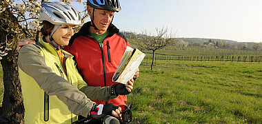 Ein Mann und eine Frau mit Radhelmen und Fahrrad stehen mit einer Karte vor einer Wiese