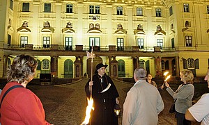 Ein Mann in historischem Nachtwächterkostüm vor einem Schloss und drei Personen mit Fackeln