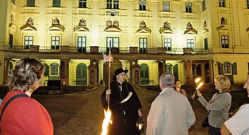 Ein Mann in historischem Nachtwächterkostüm vor einem Schloss und drei Personen mit Fackeln