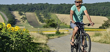Ein Mann mit grünem T-Shit und Strohhut fährt mit einem Rad durch Felder