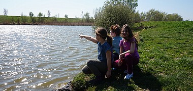 Drei Kinder sitzen in der Wiese am Ufer eines Baches.