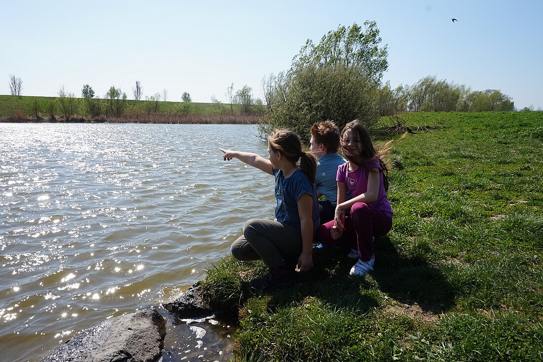 Drei Kinder sitzen in der Wiese am Ufer eines Baches.