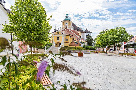 Begrünter Platz mit gelber Barockkirche mit großem Stiegenaufgang.