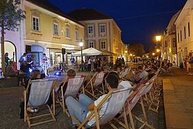 Menschen sitzen in Liegestühlen auf einem Platz, wo eine Live-Band spielt.