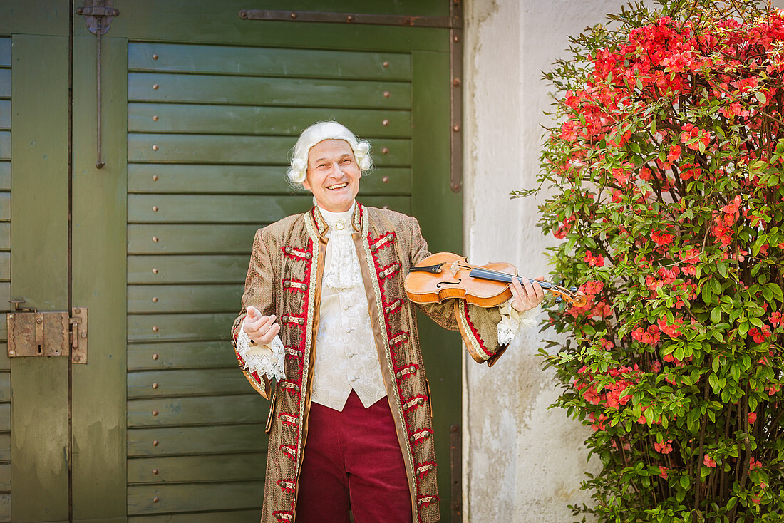 Mann in barockem Kostüm mit Geige vor einem grünen Tor und Rosenstrauch