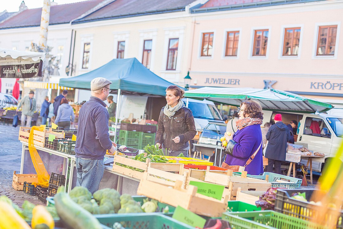 Zwei Frauen stehen vor einem Marktstand mit frischem Gemüse und einem Verkäufer