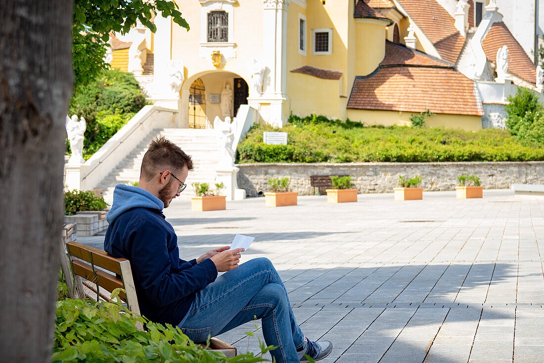 Ein Mann sitzt lesend auf einer Bank vor einen großen Kirche unter einem Baum.