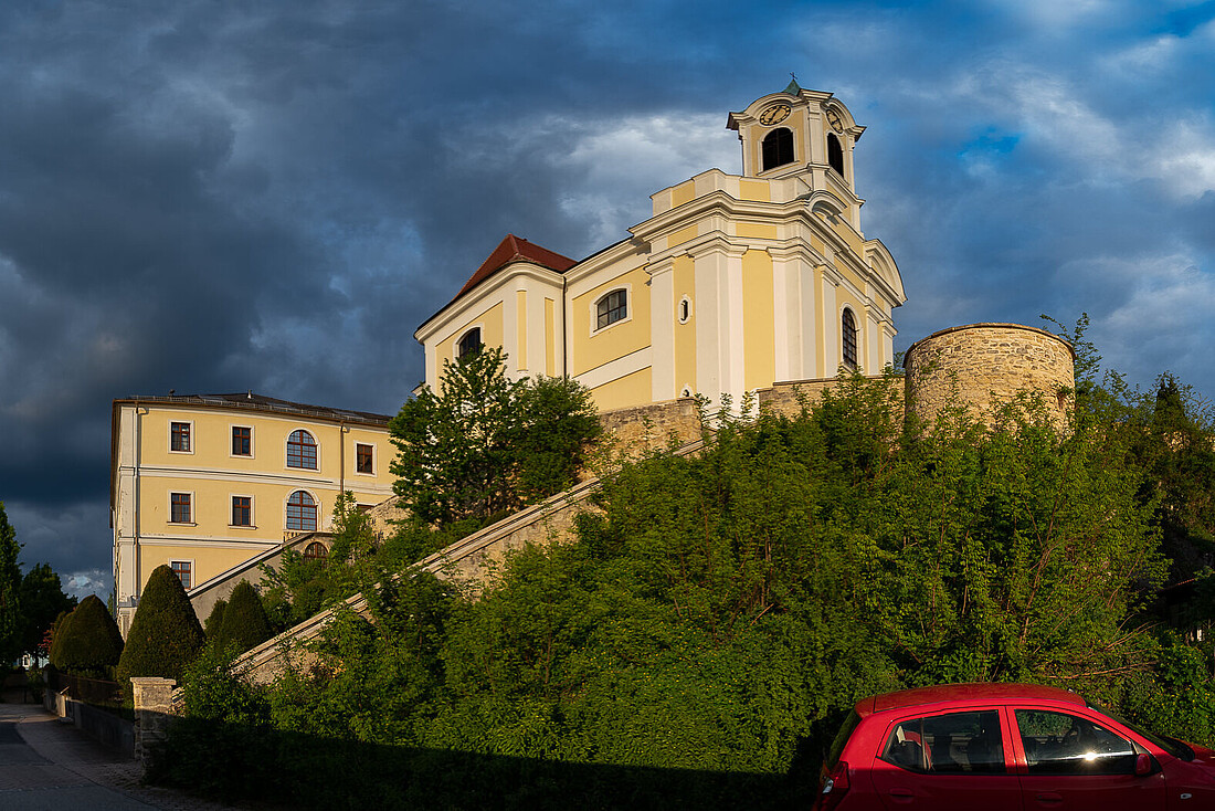 Gelbe barocke Kirche und ein Nebengebäude auf einer Anhöhe.