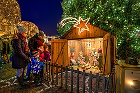 Zwei Kinder und zwei Erwachsene stehen vor einer großen Weihnachtskrippe im Freien unter einem Weihnachtsbaum