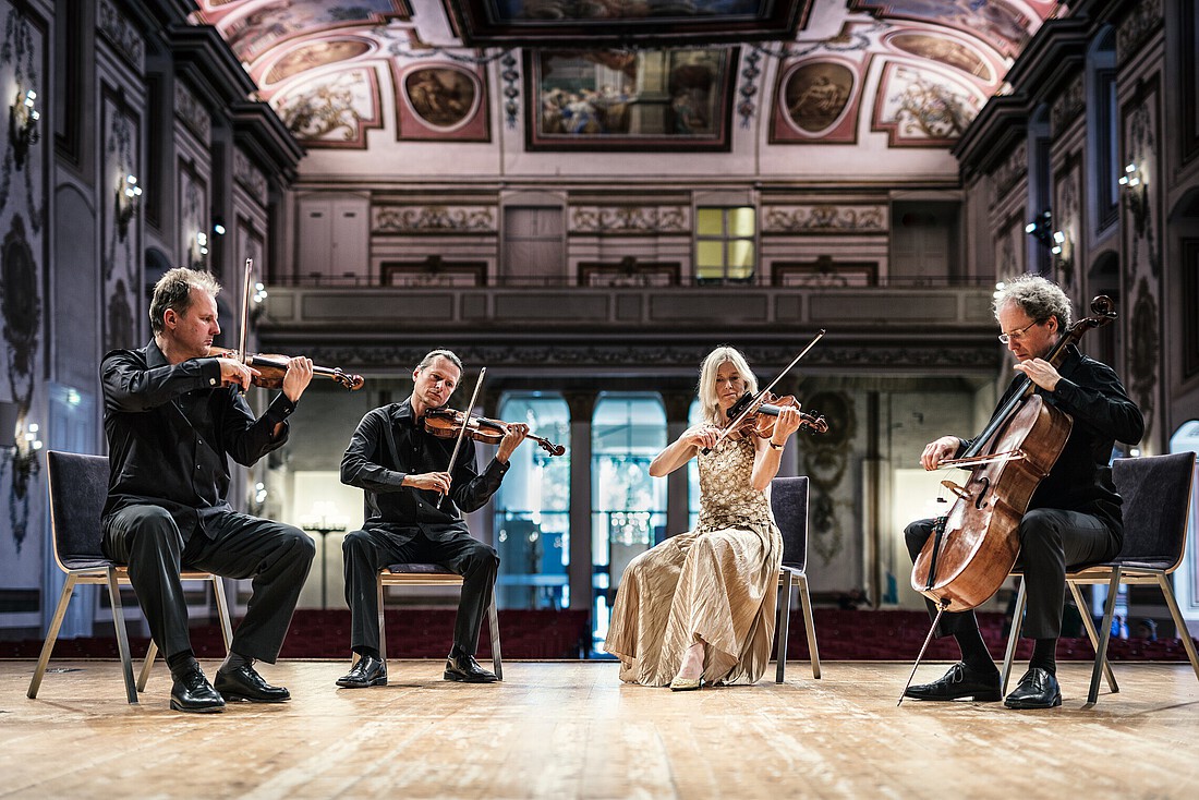 Drei Männer und eine Frau als Streichquartett in einem großen, barocken Konzertsaal.