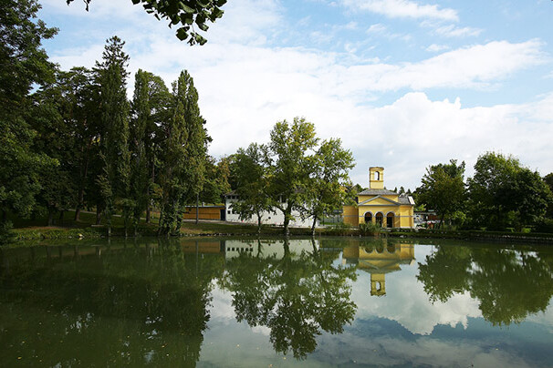 Grüner Teich mit historischem, gelben Gebäude und Bäumen am Ufer.