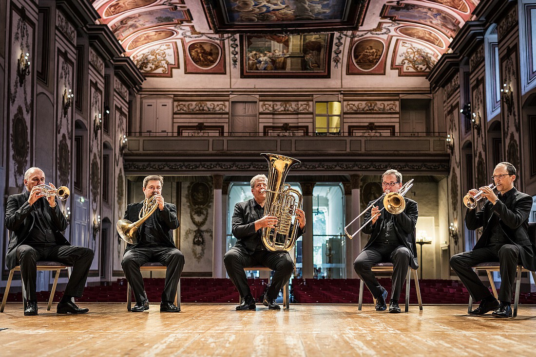 Fünf Männer mit Blechblasinstrumenten auf einer Bühne in einem großen barocken Konzertsaal.