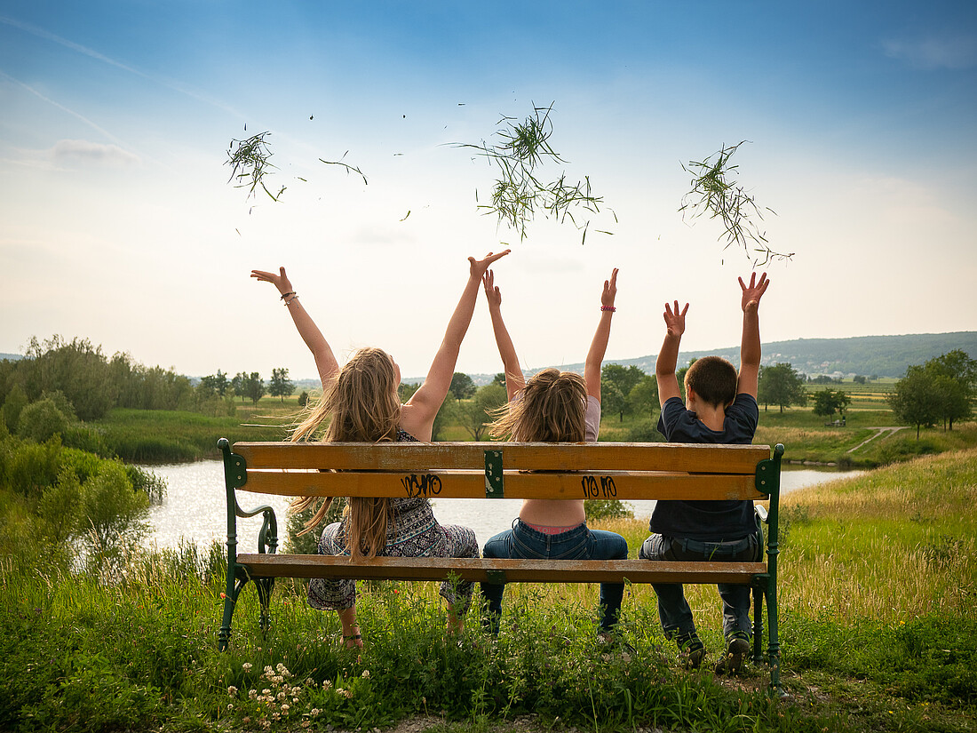 Drei Mädchen sitzen auf einer Bank bei einem Teich und werfen Grashalme in die Luft.