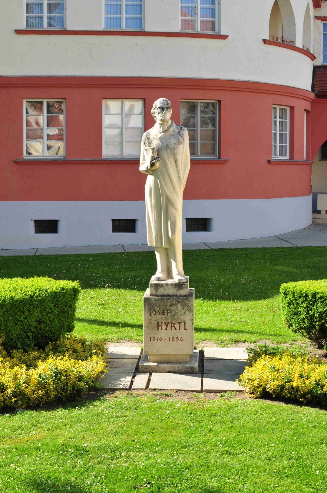 Männliche Person als Denkmal umgeben von einer Wiese, vor rotem Haus.